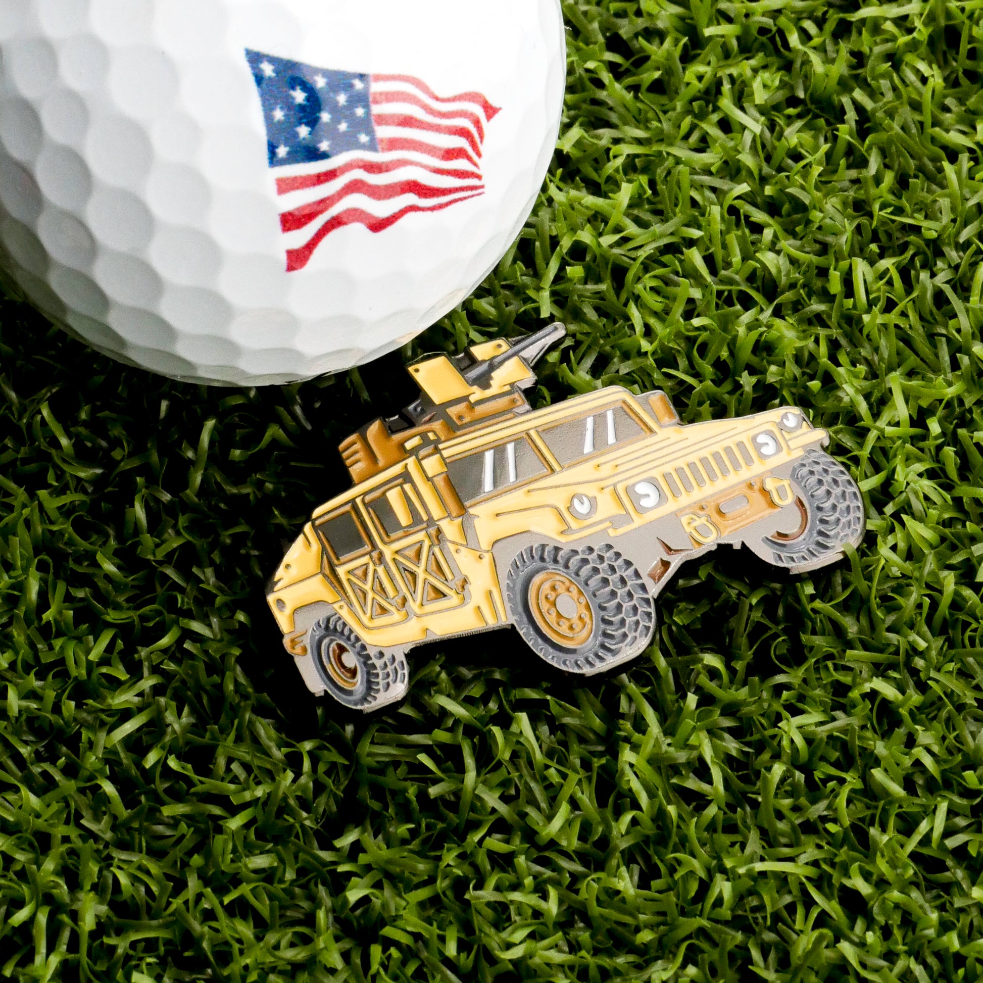 A close-up of the Humvee Ball Marker by Ballmarkers.com, featuring an American flag on a golf ball beside a yellow toy military vehicle on green grass, brings bold tactical style to your game.
