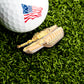 A golf ball with an American flag graphic rests on grass beside the Ballmarkers.com Tank Ball Marker, creating a unique military-themed golf accessory.