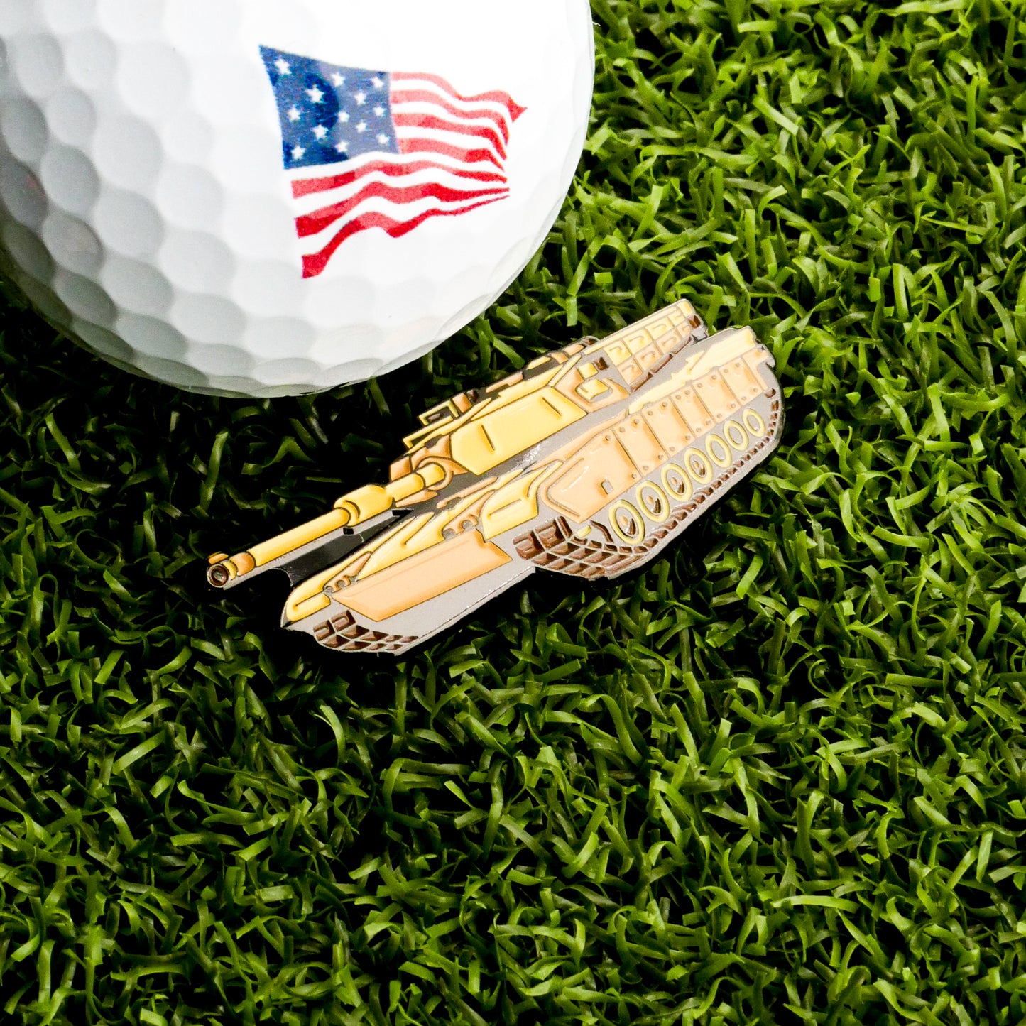 A golf ball with an American flag graphic rests on grass beside the Ballmarkers.com Tank Ball Marker, creating a unique military-themed golf accessory.