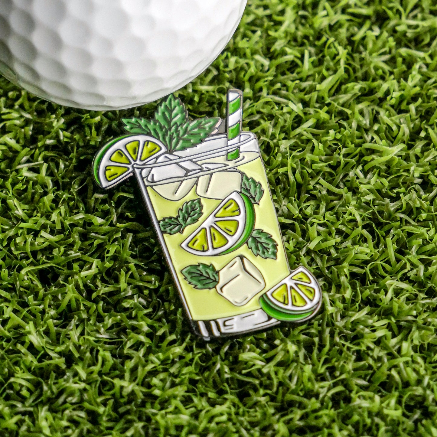 The Mojito Ball Marker from Ballmarkers.com features a glass with lime, mint, ice, and a striped straw resting on grass beside a white golf ball—perfect for adding cocktail flair to your collection of golf accessories.
