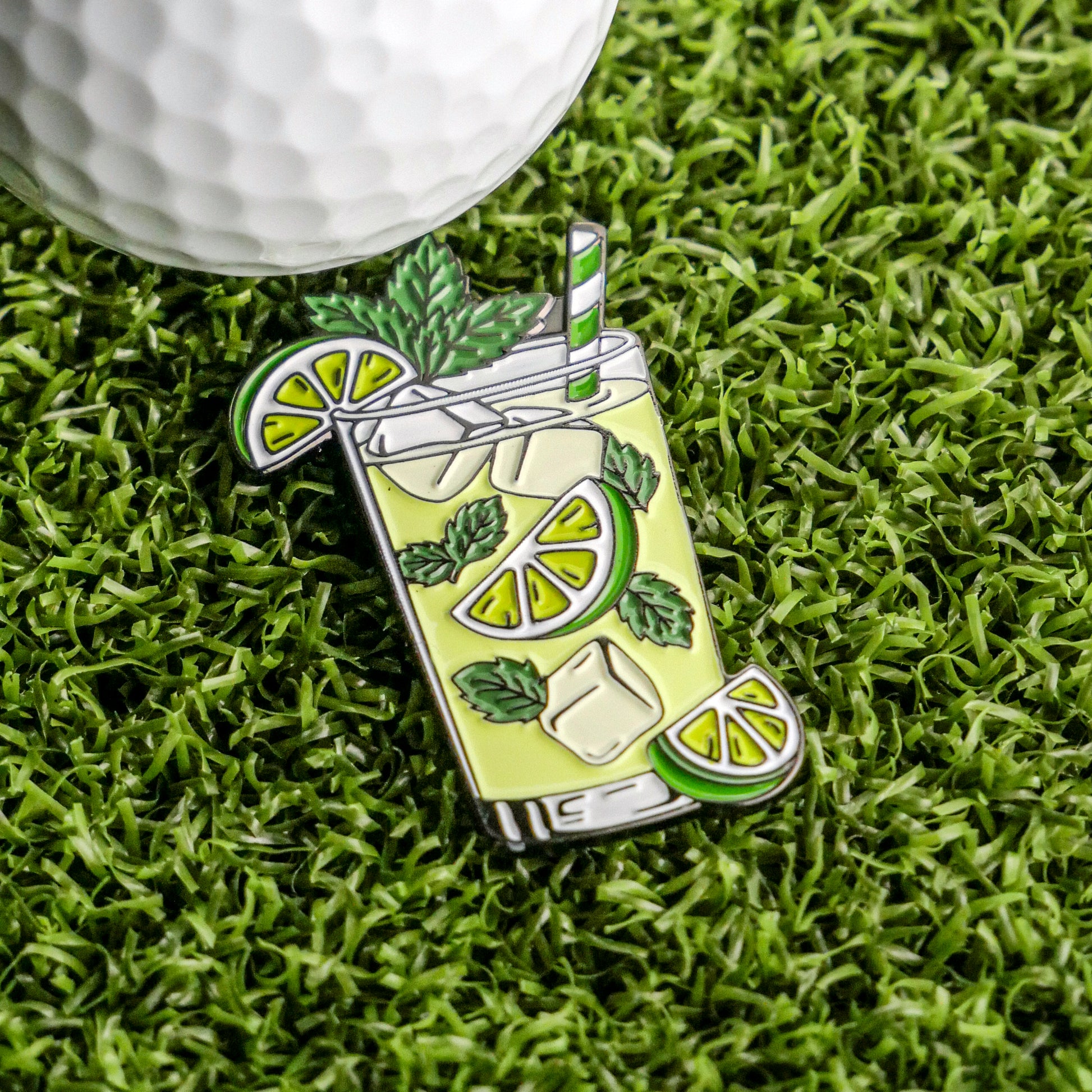 The Mojito Ball Marker from Ballmarkers.com features a glass with lime, mint, ice, and a striped straw resting on grass beside a white golf ball—perfect for adding cocktail flair to your collection of golf accessories.