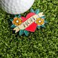 The Mom Tattoo Ball Marker by Ballmarkers.com features "MOM" on a heart with yellow flowers and leaves, inspired by mom tattoo designs. Shown on artificial grass next to a golf ball, it makes a unique Mother’s Day gift for golfers.