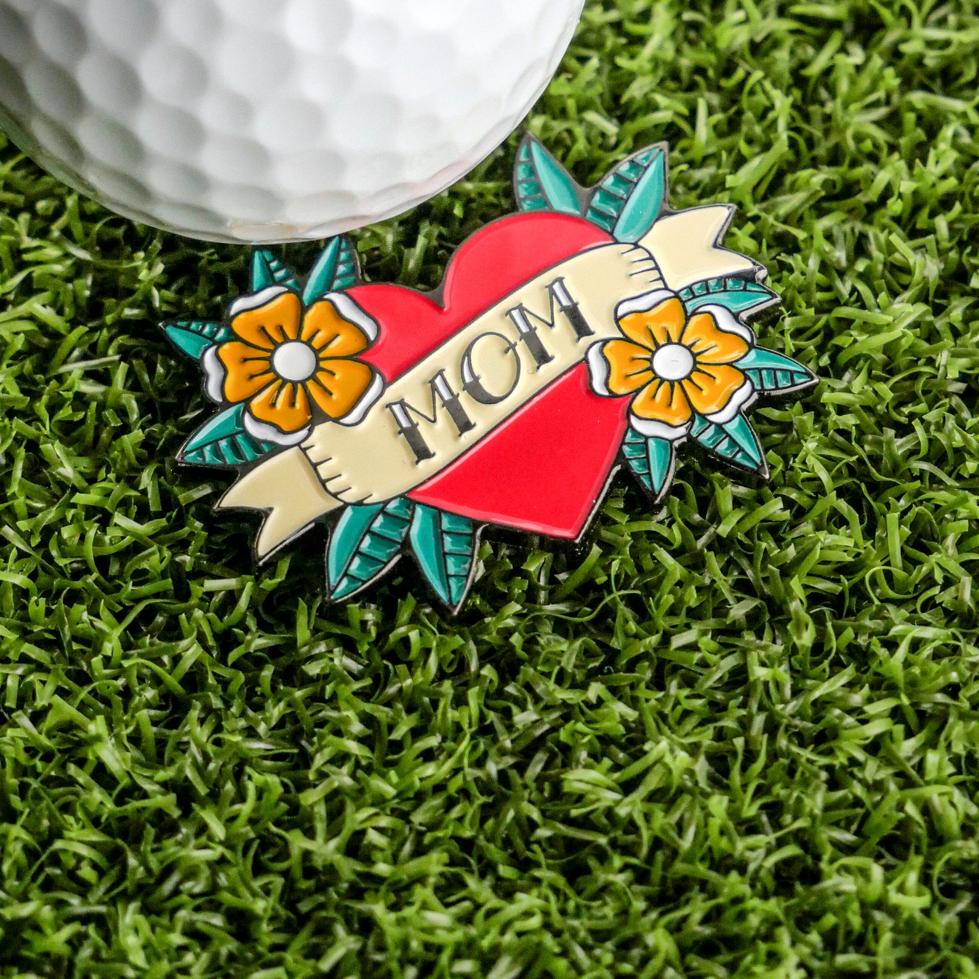 The Mom Tattoo Ball Marker by Ballmarkers.com features "MOM" on a heart with yellow flowers and leaves, inspired by mom tattoo designs. Shown on artificial grass next to a golf ball, it makes a unique Mother’s Day gift for golfers.