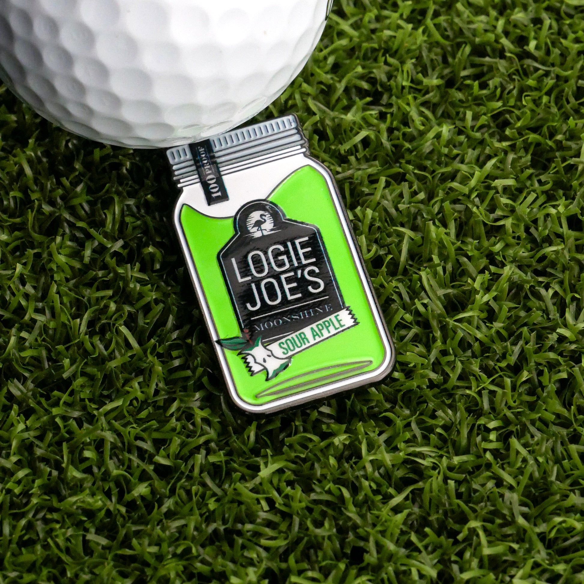 A close-up of a golf ball next to the Logie Joe's Golf Moonshine Ball Marker on bright green artificial grass, highlighting fun and unique golf accessories.