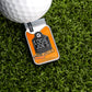 A golf ball on artificial grass sits next to the Moonshine Ball Marker by Logie Joe's Golf, designed like an orange mason jar labeled Logie Joe’s Moonshine Apple Pie with a pie slice illustration—a unique golf accessory.