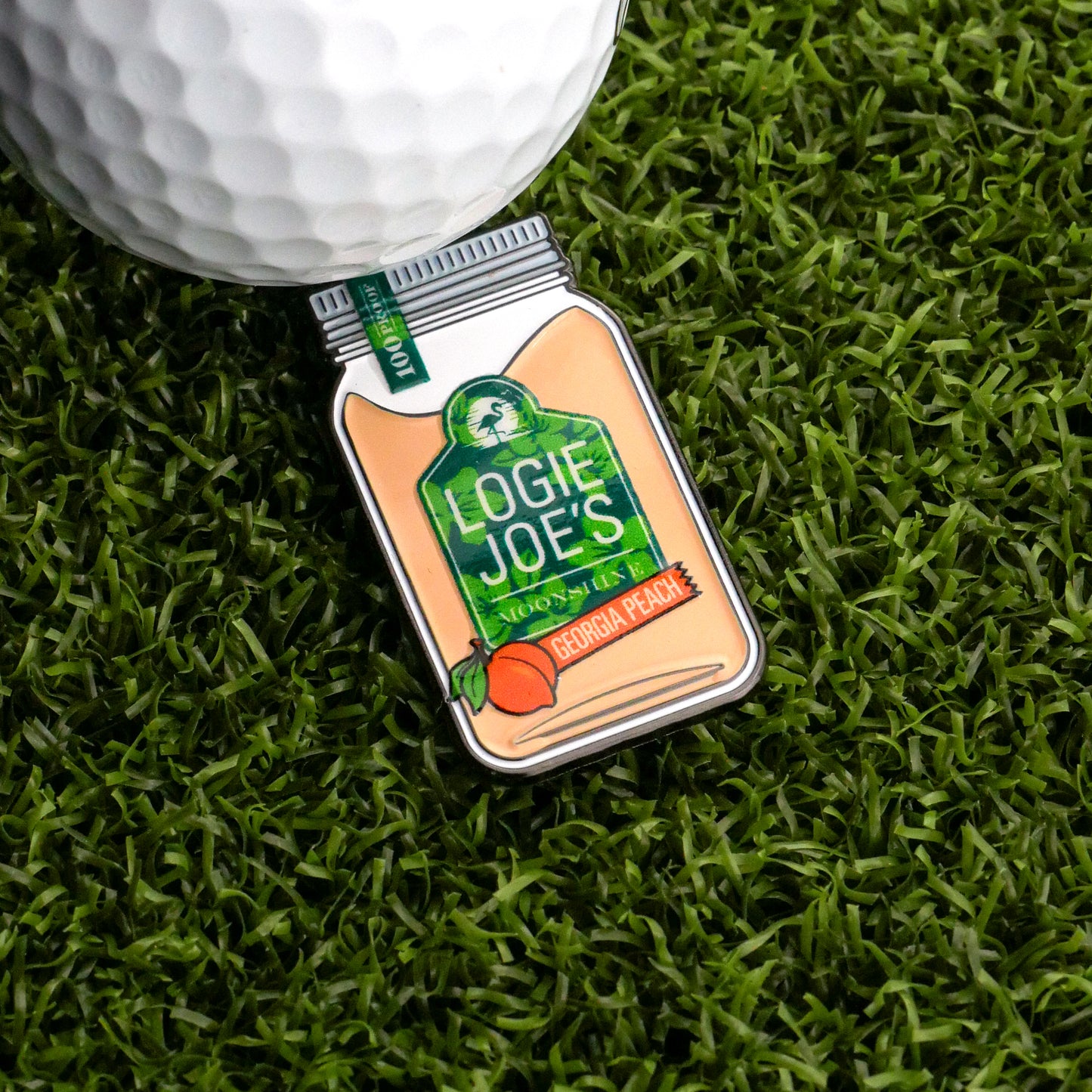 A golf ball sits on artificial grass beside a mason jar-shaped Moonshine Ball Marker by Logie Joe's Golf, labeled "Georgia Peach," featuring a peach illustration and a green lid.