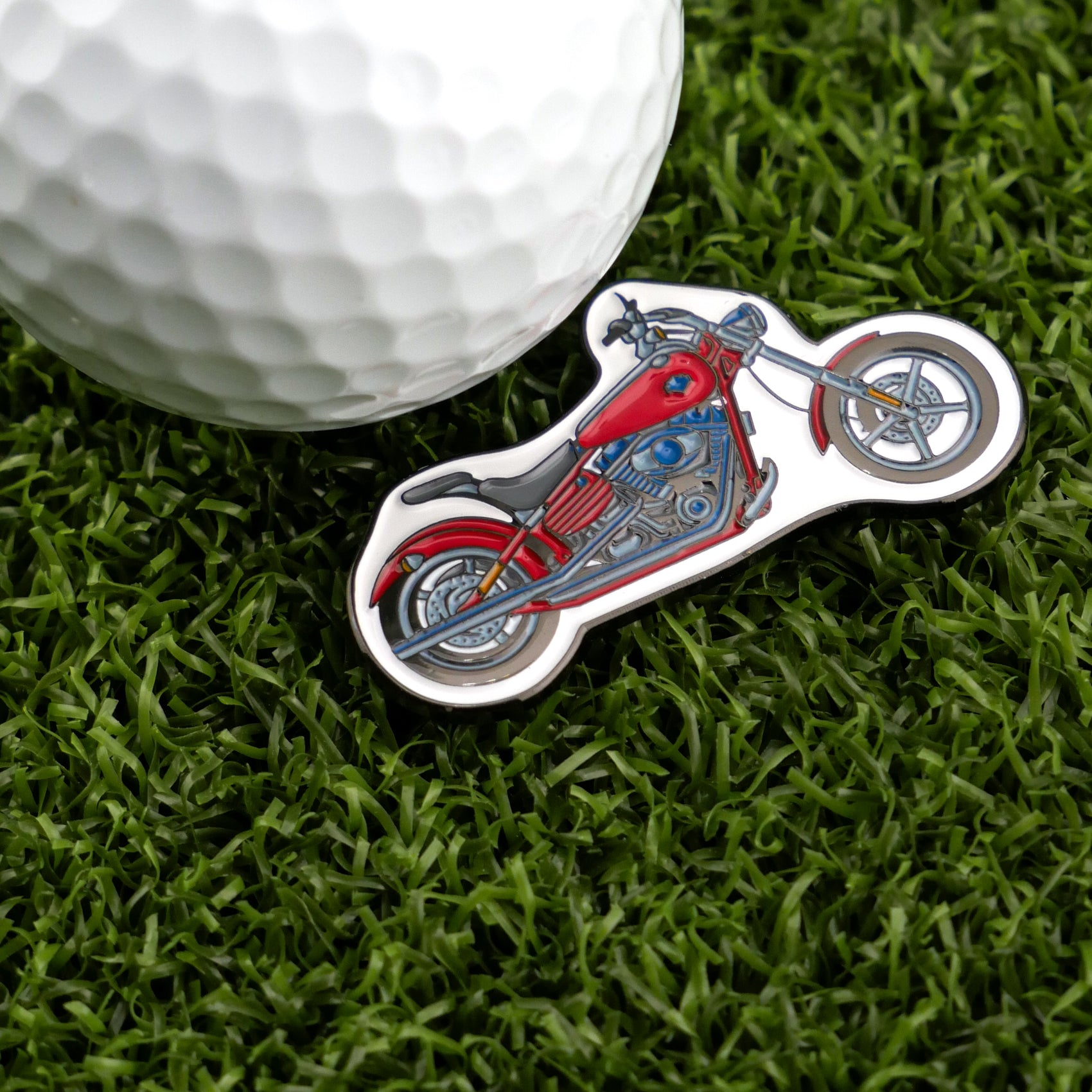 A Logie Joe's Golf white ball rests on green grass beside the Motorcycle pin-shaped red ball marker, adding flair to your premium golf accessories collection.