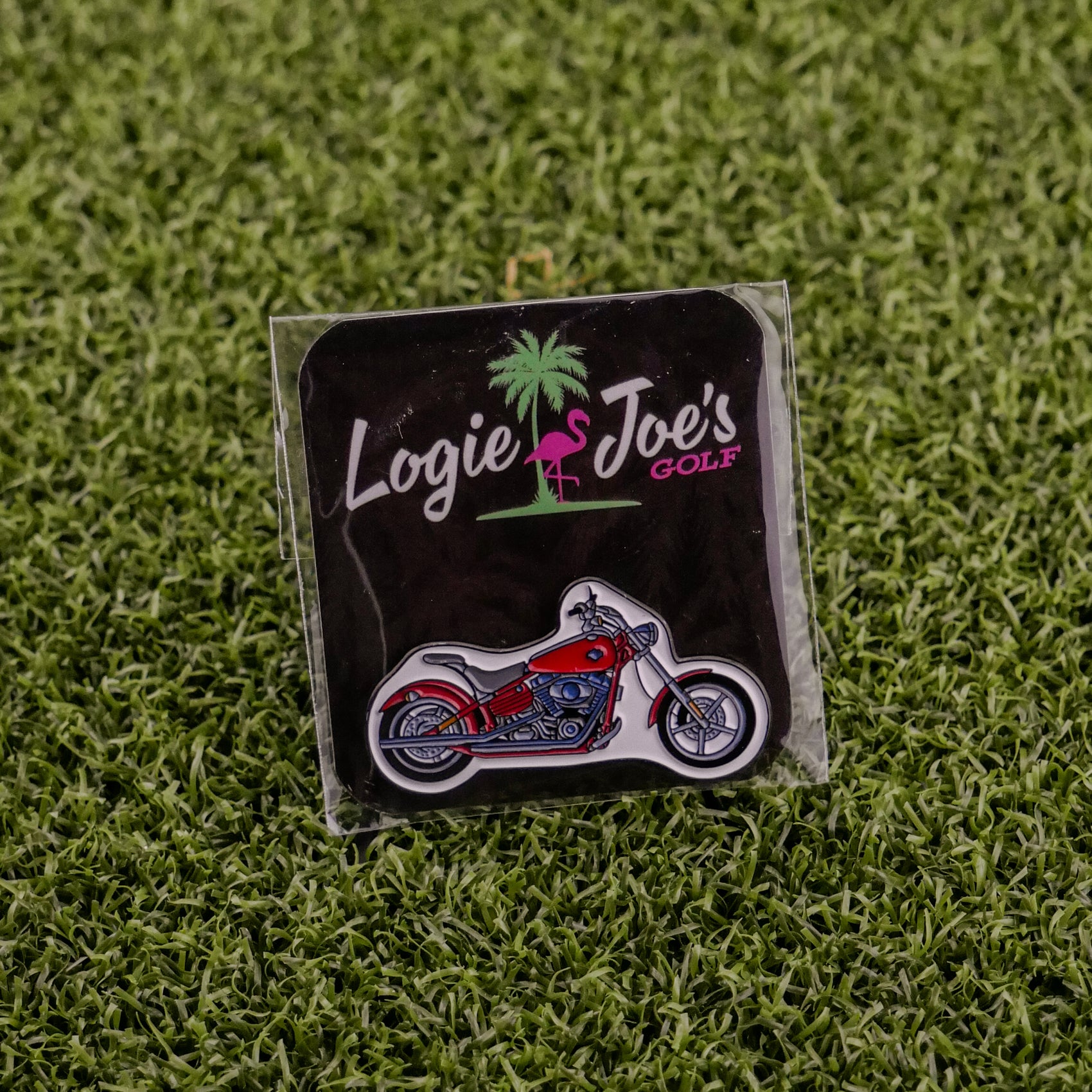 A red and silver Motorcycle ball marker pin by Logie Joe’s Golf, presented on a black card with colorful branding and set on green artificial grass—a standout addition to premium golf accessories.