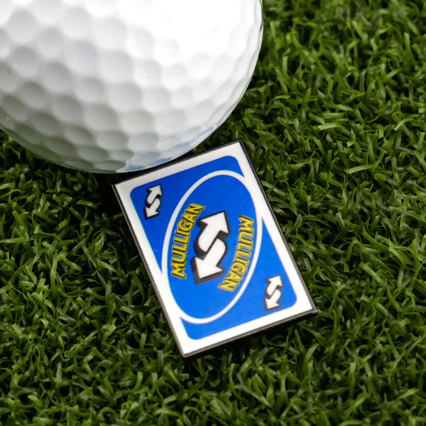 A golf ball sits on artificial grass beside Logie Joe's Golf Mulligan Reverse Card Blue—a fun UNO-style marker with "MULLIGAN" in yellow on blue, ideal for fans of unique golf ball markers and premium accessories.