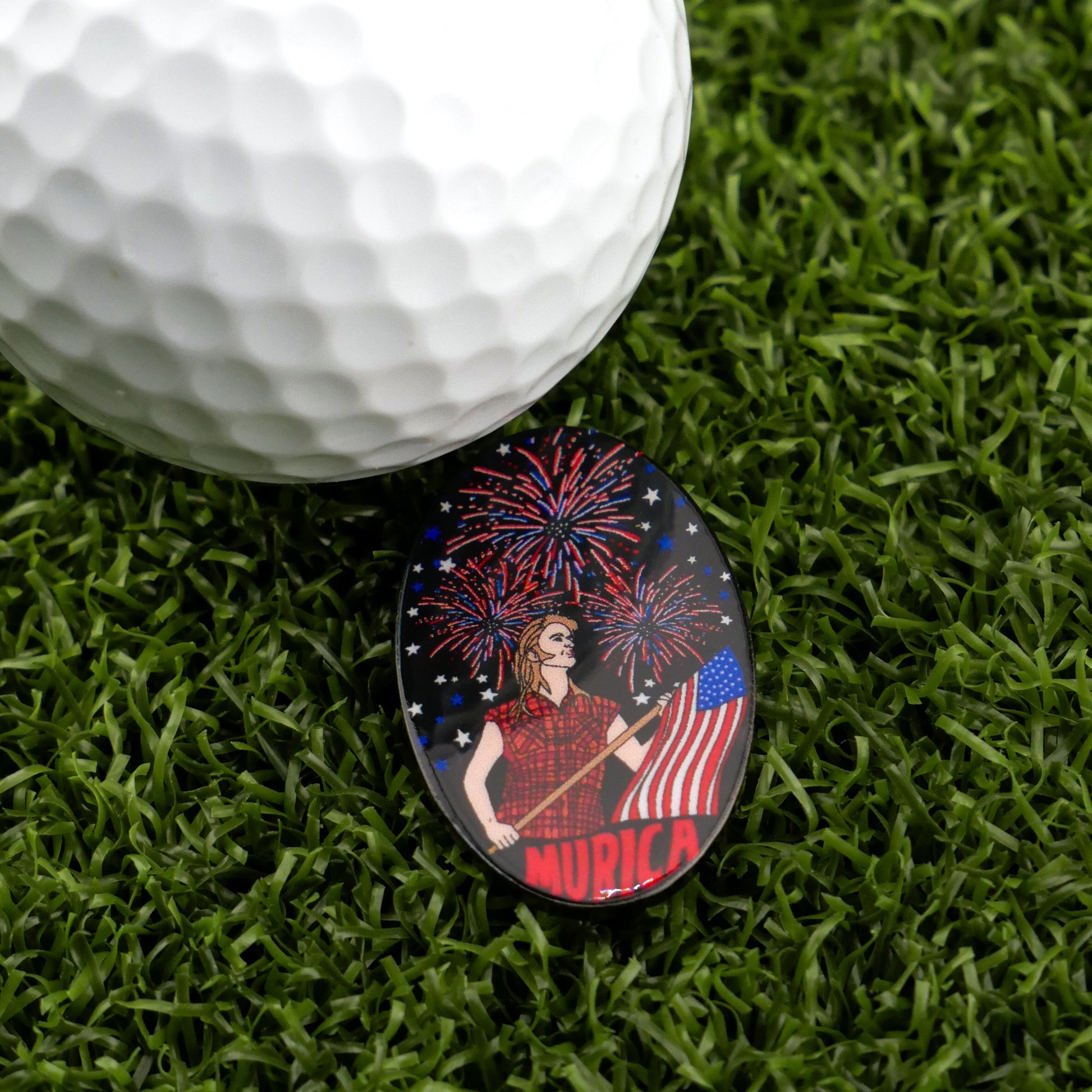 A close-up of a golf ball on artificial grass next to Logie Joe's Golf "Murica" ball marker—featuring a flag-waving figure, fireworks, and ‘MURICA’ text—a funny patriotic accessory for golfers.