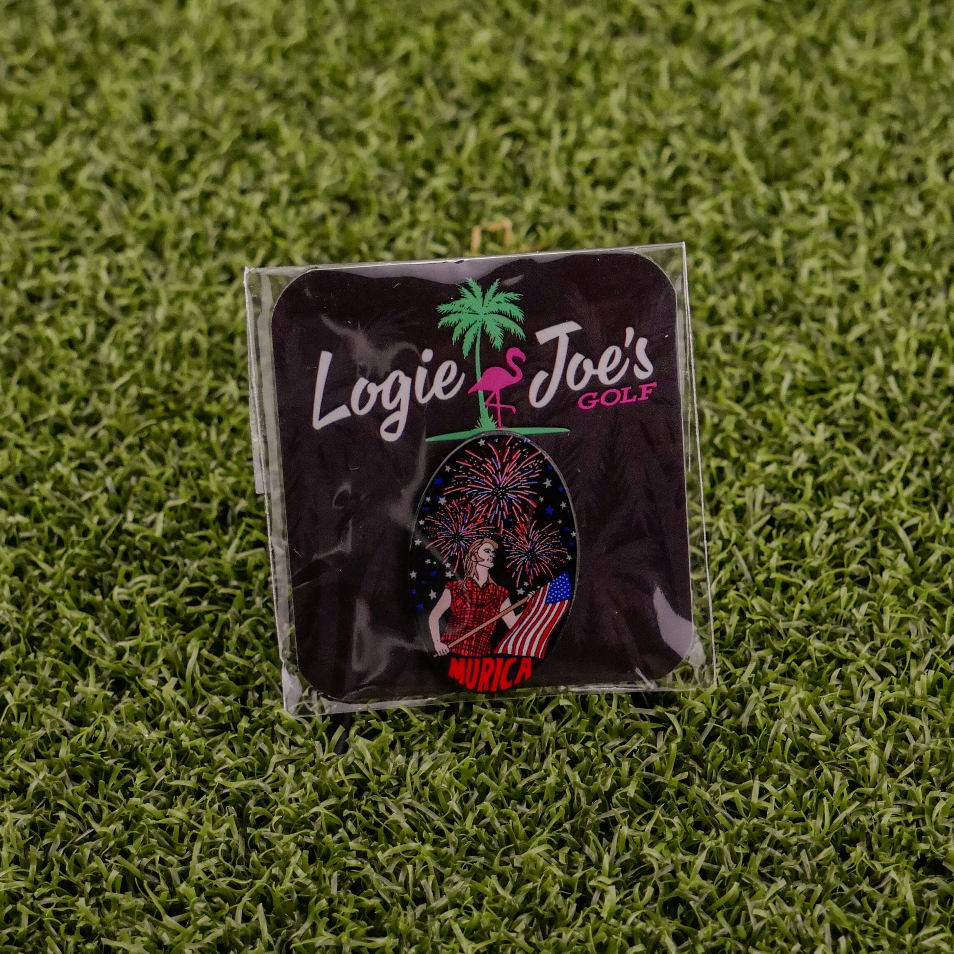 A square sticker on artificial grass reads "Logie Joe's Golf" with a palm tree and flamingo. Below, a woman holds an American flag amid fireworks and "Murica"—a funny Logie Joe's Golf premium ball marker accessory.