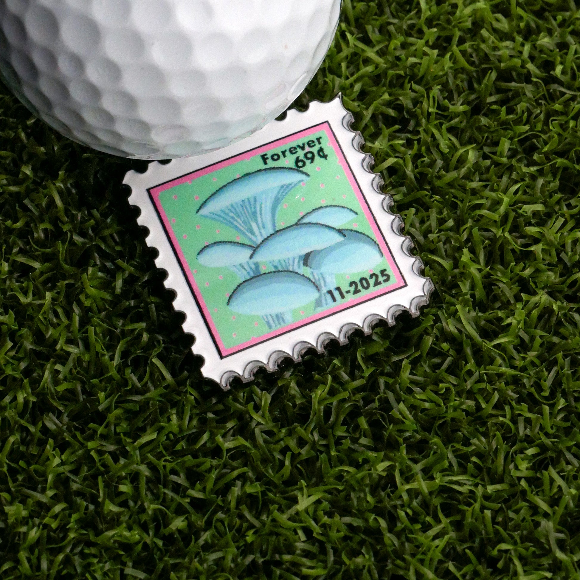 A November Mushroom Stamp Ball Marker by Ballmarkers.com rests on artificial grass beside a white golf ball—an ideal addition for unique stamp collectors.