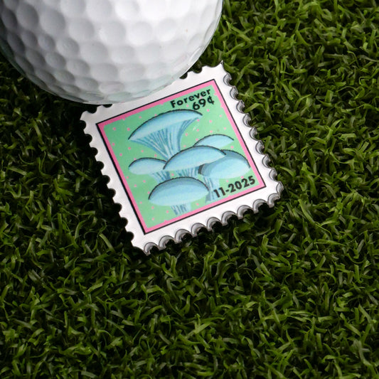 A November Mushroom Stamp Ball Marker by Ballmarkers.com rests on artificial grass beside a white golf ball—an ideal addition for unique stamp collectors.