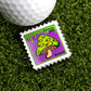 The 10-2025 Mushroom Stamp Ball Marker by Ballmarkers.com features a yellow and green mushroom with a brown stem on purple, reading "Forever 99¢"—a unique golf accessory beside your ball on the green.