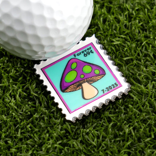A golf ball sits on artificial grass next to Logie Joe's Golf Mushroom Stamp 07/2025 Ball Marker, shaped like a stamp with a cartoon purple and green mushroom, 