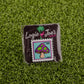 The Mushroom Stamp 07/2025 by Logie Joe's Golf is a colorful enamel ball marker shaped like a postage stamp, featuring a purple and green mushroom with “Forever 69¢ 7-2025,” displayed on grass with branded backing.