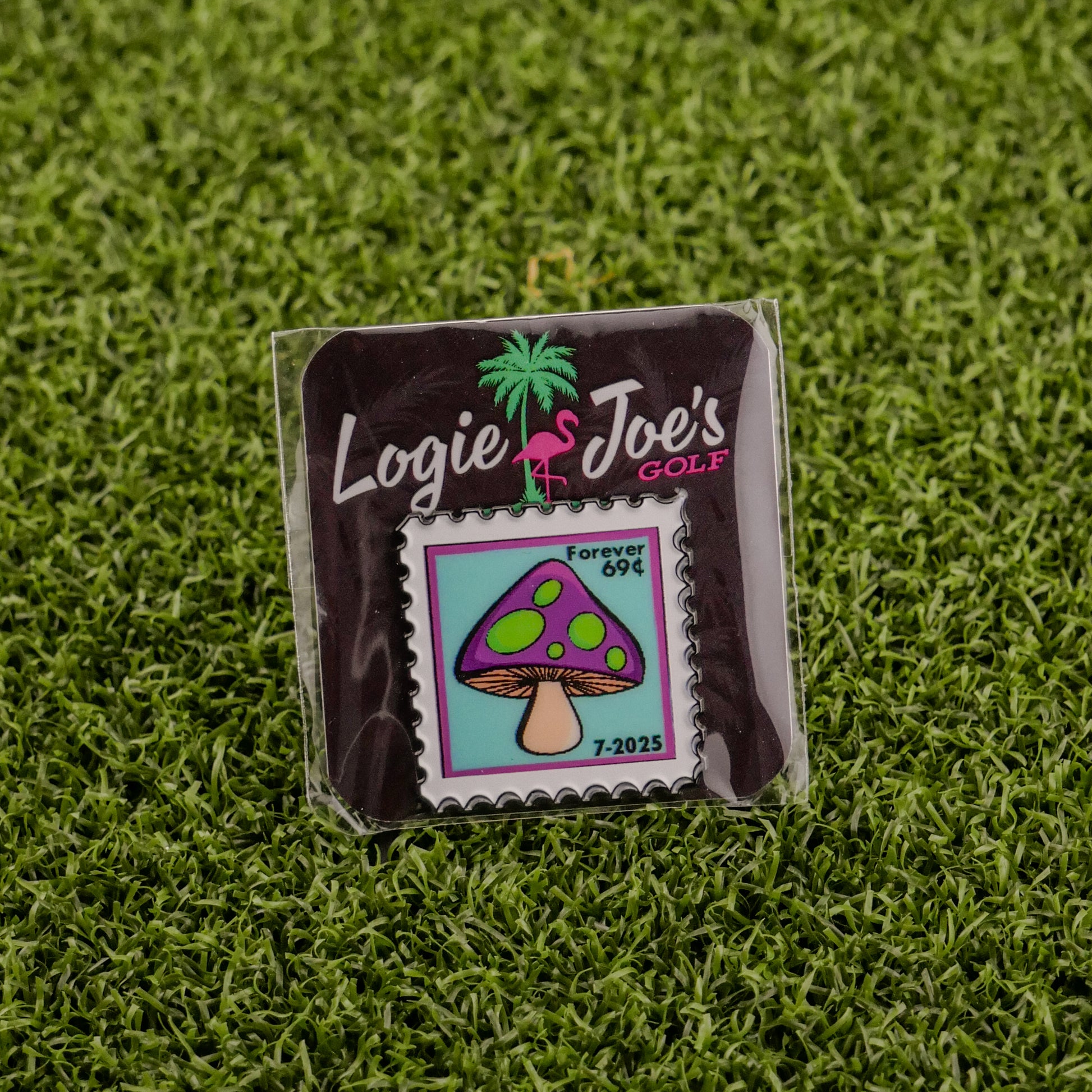 The Mushroom Stamp 07/2025 by Logie Joe's Golf is a colorful enamel ball marker shaped like a postage stamp, featuring a purple and green mushroom with “Forever 69¢ 7-2025,” displayed on grass with branded backing.