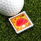 A close-up of a golf ball on artificial grass next to Logie Joe's Golf Mushroom Stamp 08/2025 marker, featuring a red and white mushroom on yellow and designed like a postage stamp.