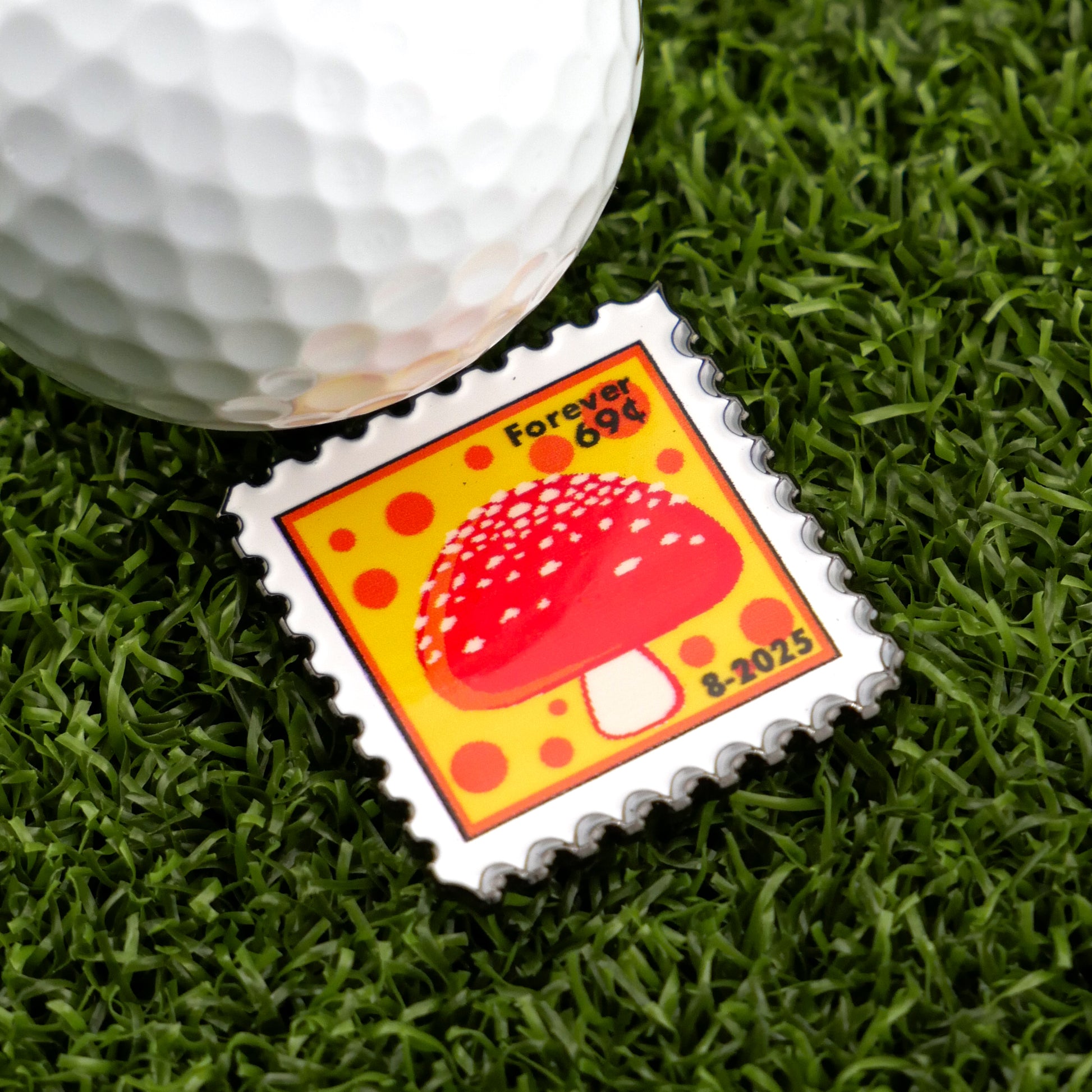 A close-up of a golf ball on artificial grass next to Logie Joe's Golf Mushroom Stamp 08/2025 marker, featuring a red and white mushroom on yellow and designed like a postage stamp.