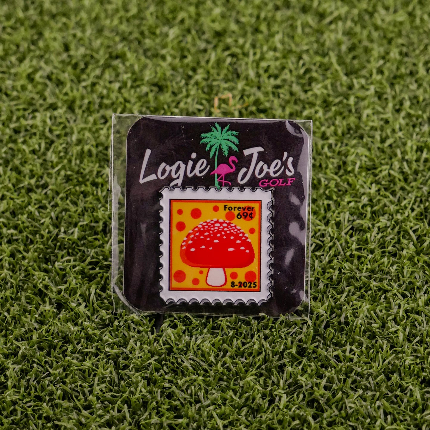 The Mushroom Stamp 08/2025 by Logie Joe’s Golf is a square enamel ball marker, styled like a postage stamp with a red mushroom and yellow background, marked “Forever 69¢, 8-2025,” packaged on brand card atop green artificial grass.