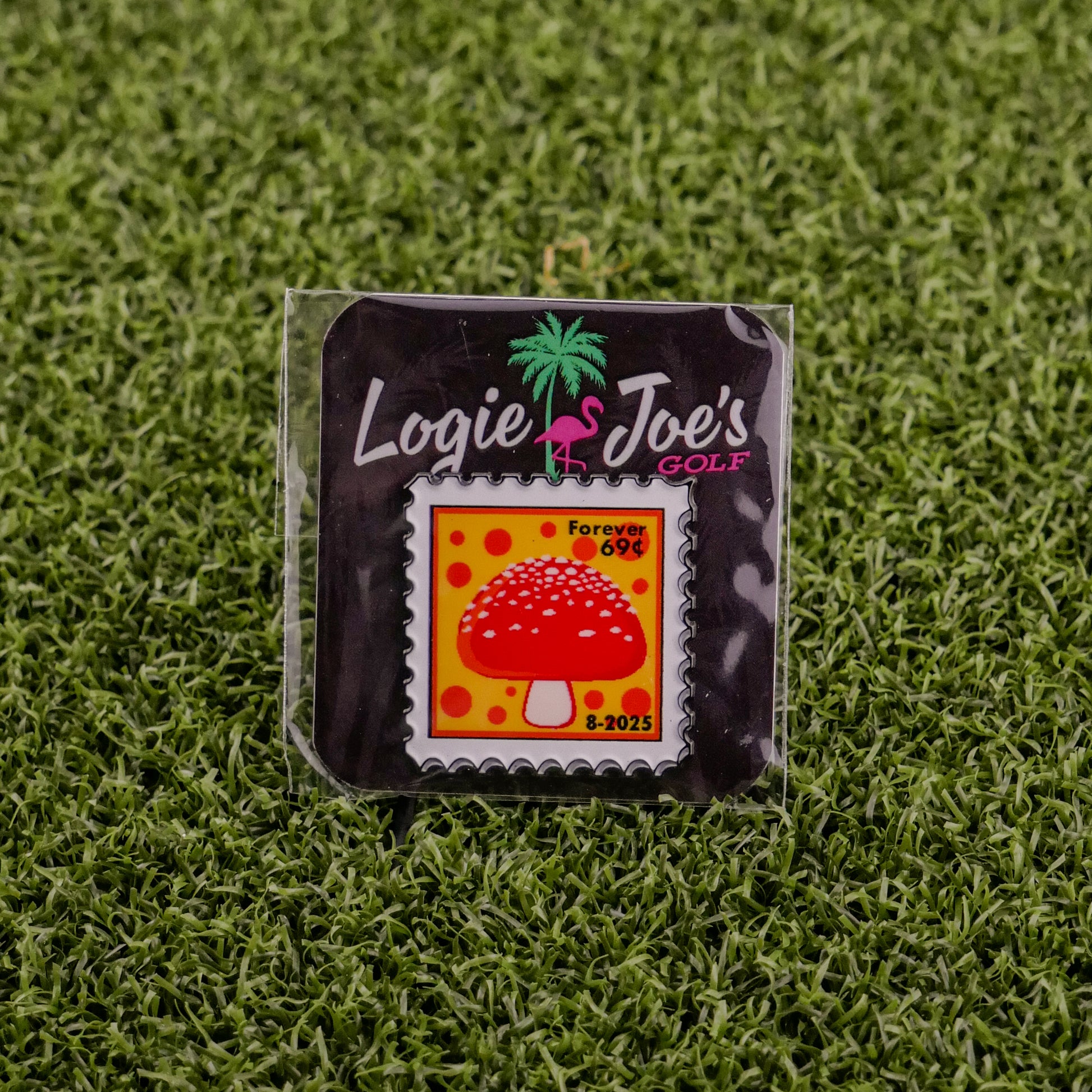 The Mushroom Stamp 08/2025 by Logie Joe’s Golf is a square enamel ball marker, styled like a postage stamp with a red mushroom and yellow background, marked “Forever 69¢, 8-2025,” packaged on brand card atop green artificial grass.