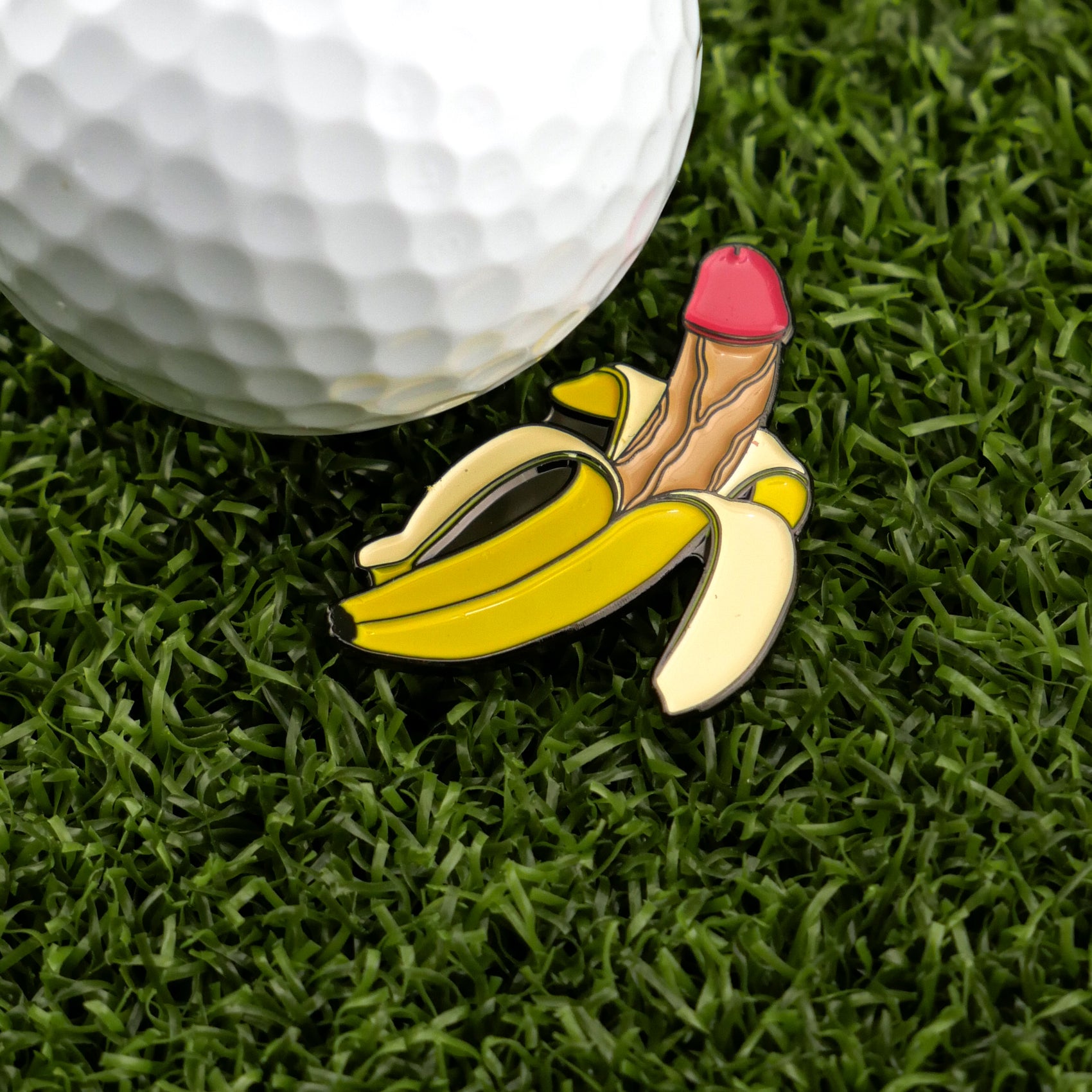 A golf ball sits on green grass next to Logie Joe's Golf Nannerdick, a novelty banana-shaped premium ball marker humorously resembling a penis—perfect for golfers who enjoy playful, artistic accessories.