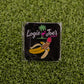 A Nannerdick pin by Logie Joe's Golf, shaped like a peeled banana with a pink tip, rests on grass. Behind, a black card displays the Logie Joe's Golf logo—a pink flamingo and palm—making this an eye-catching golf ball marker.