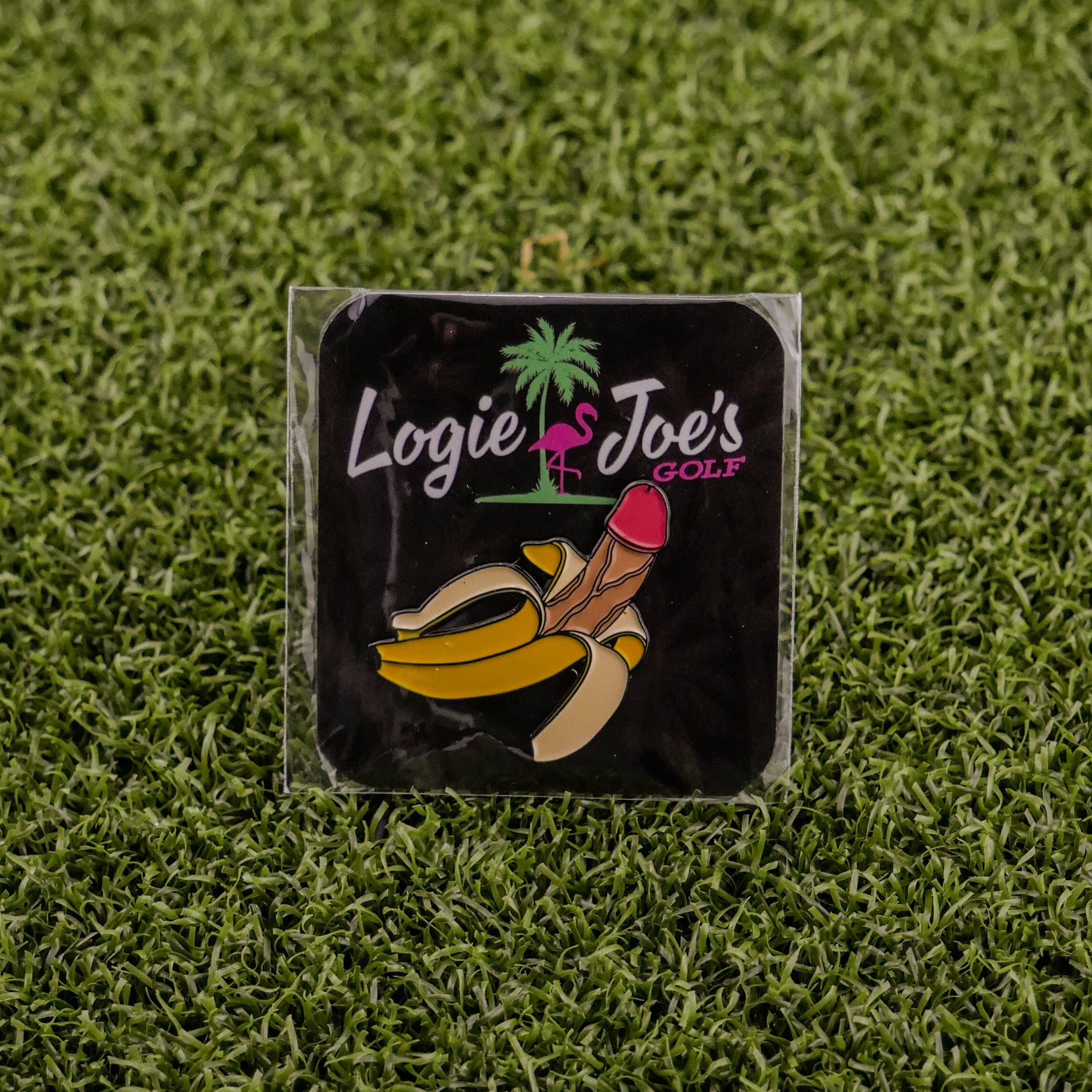A Nannerdick pin by Logie Joe's Golf, shaped like a peeled banana with a pink tip, rests on grass. Behind, a black card displays the Logie Joe's Golf logo—a pink flamingo and palm—making this an eye-catching golf ball marker.
