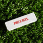 The Pins and Aces NASA "Worm" Ball Marker, a white enamel-finished rectangle with PINS & ACES. in red NASA-style font, rests on green artificial grass.