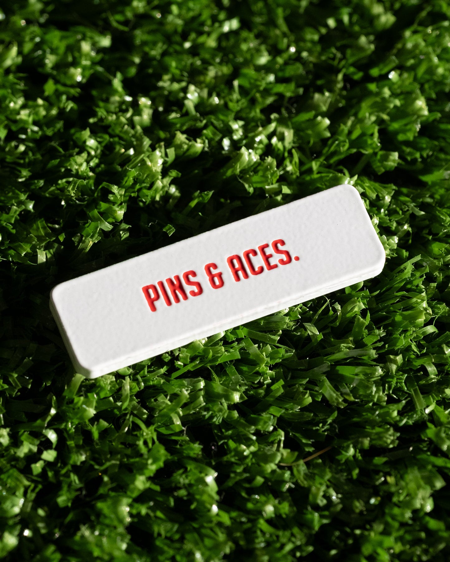 The Pins and Aces NASA "Worm" Ball Marker, a white enamel-finished rectangle with PINS & ACES. in red NASA-style font, rests on green artificial grass.