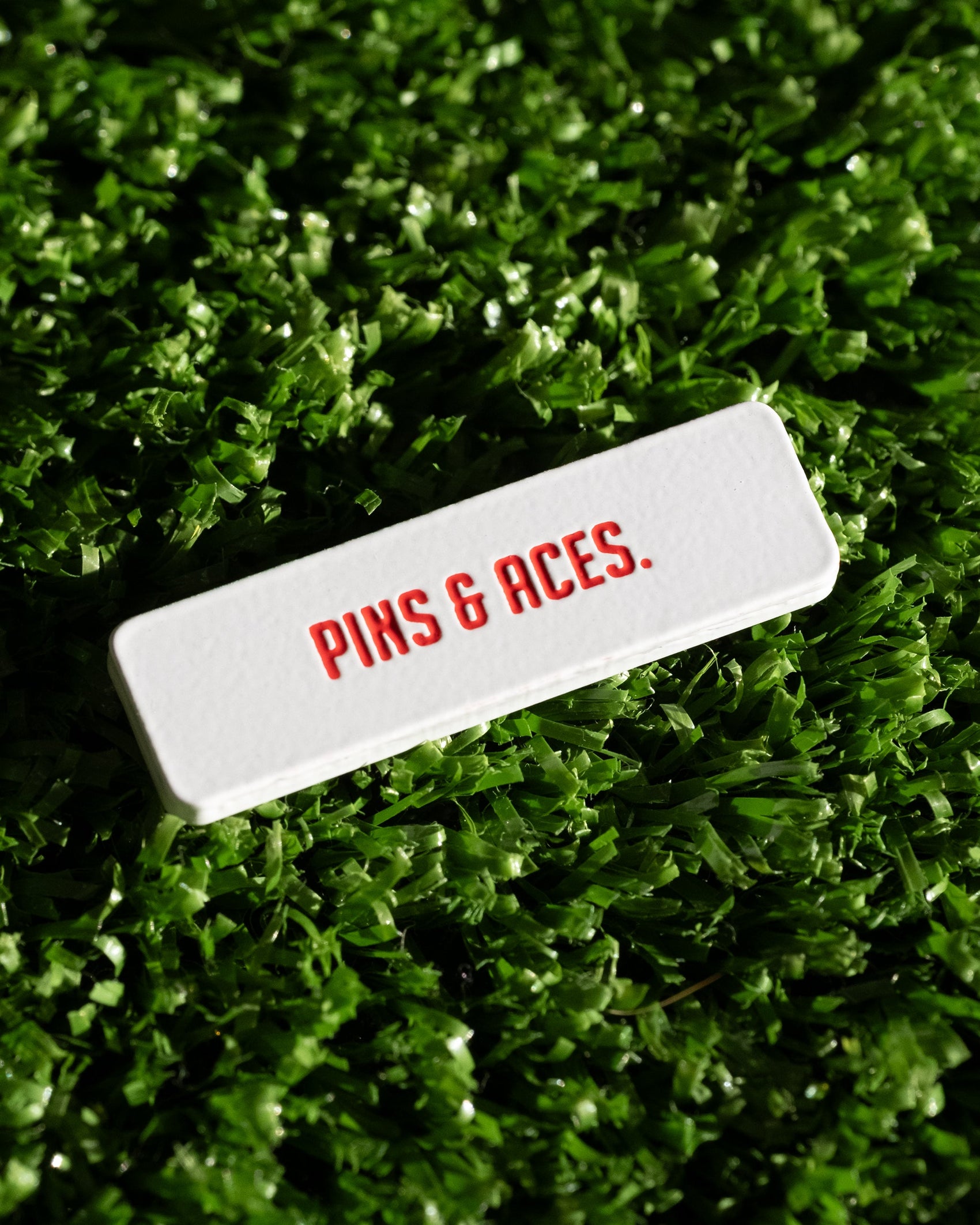The Pins and Aces NASA "Worm" Ball Marker, a white enamel-finished rectangle with PINS & ACES. in red NASA-style font, rests on green artificial grass.