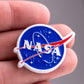A hand displays the Pins and Aces NASA "Meatball" Ball Marker, showcasing the iconic blue circular logo with white text, a red swoosh, and stars—an ideal accessory for any golf enthusiast.