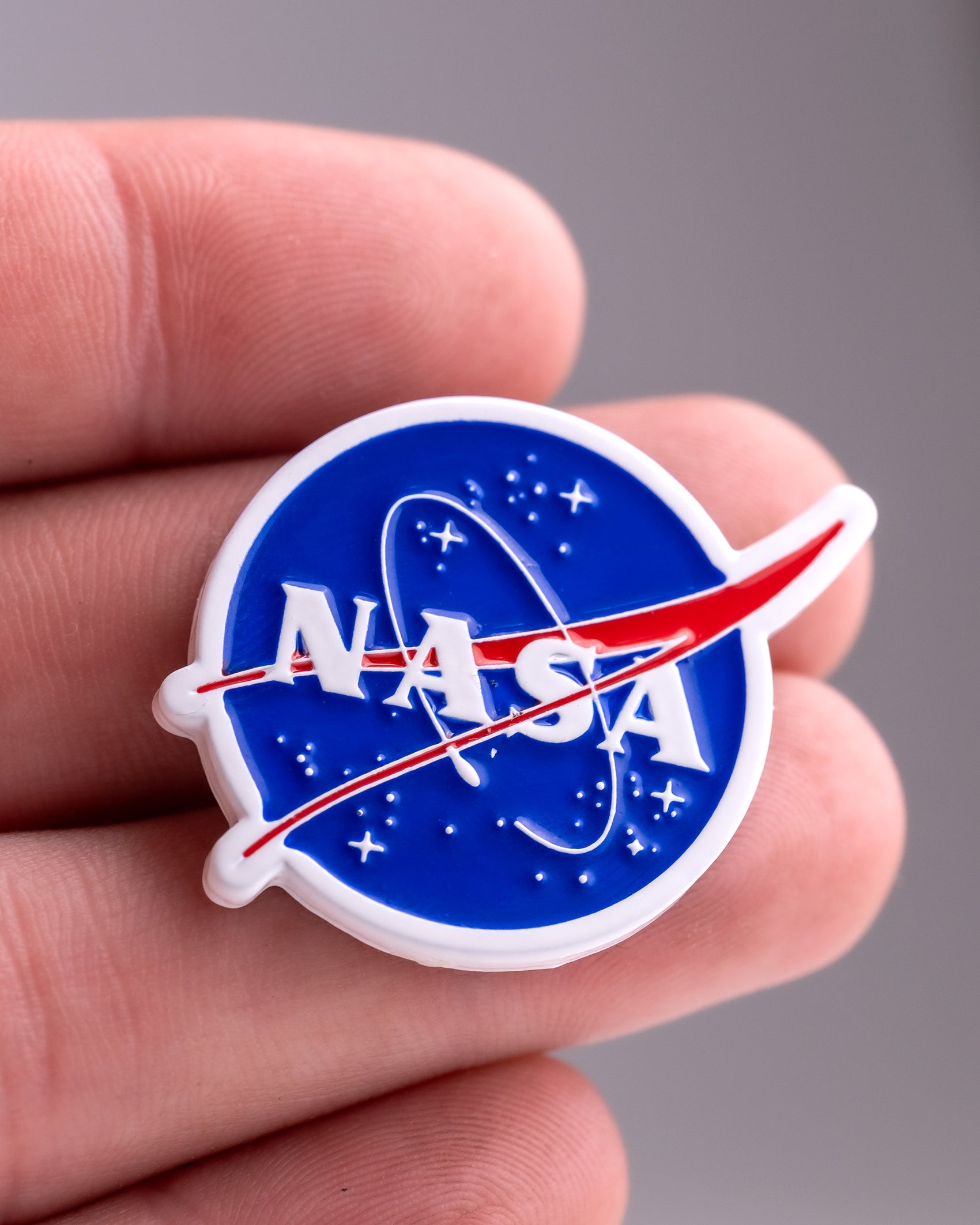 A hand displays the Pins and Aces NASA "Meatball" Ball Marker, showcasing the iconic blue circular logo with white text, a red swoosh, and stars—an ideal accessory for any golf enthusiast.