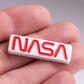 A person holds the NASA "Worm" Ball Marker by Pins and Aces, a small rectangular white marker with the bold red NASA worm logo in enamel. The blurred background emphasizes the marker and the person's fingers.