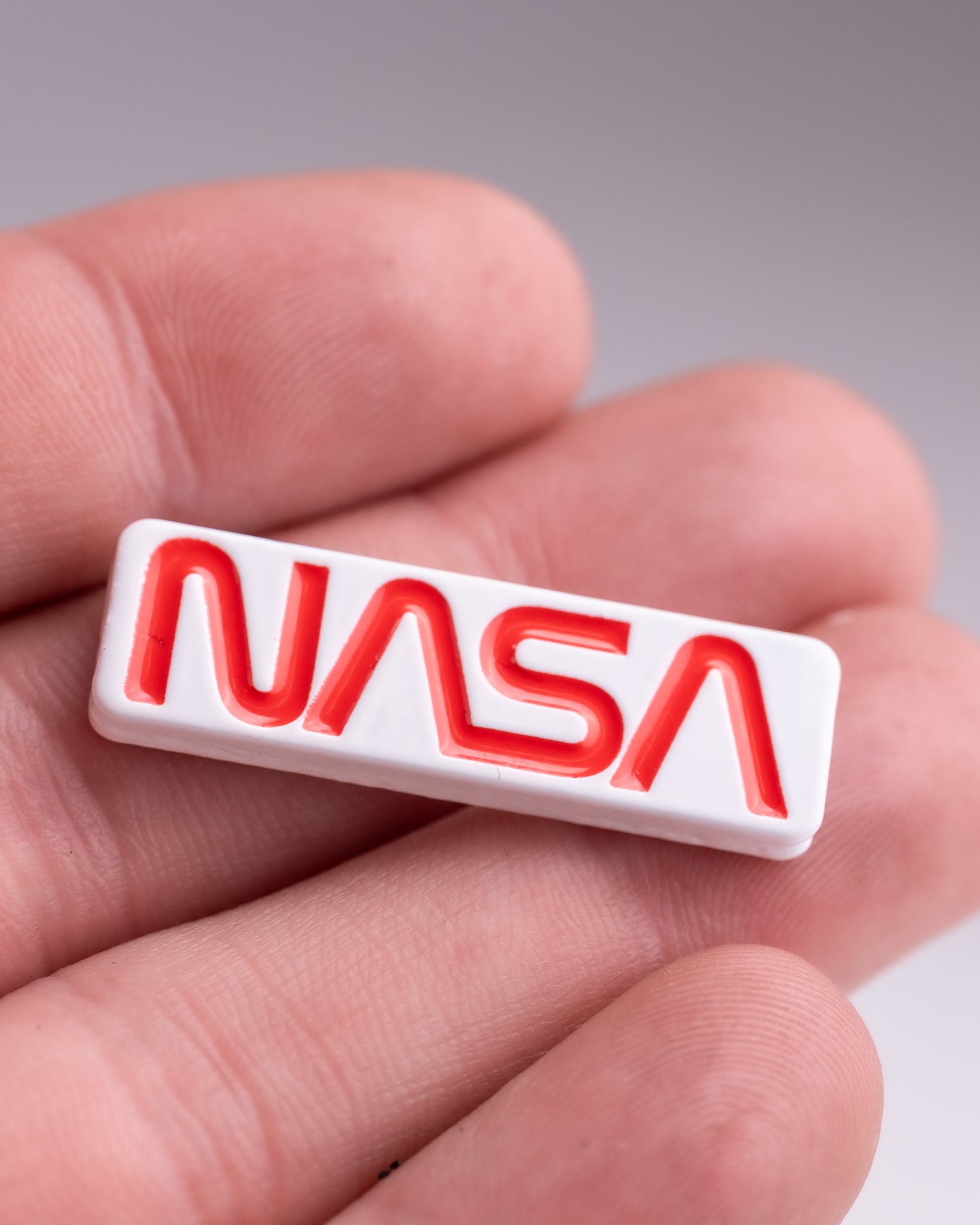 A person holds the NASA "Worm" Ball Marker by Pins and Aces, a small rectangular white marker with the bold red NASA worm logo in enamel. The blurred background emphasizes the marker and the person's fingers.