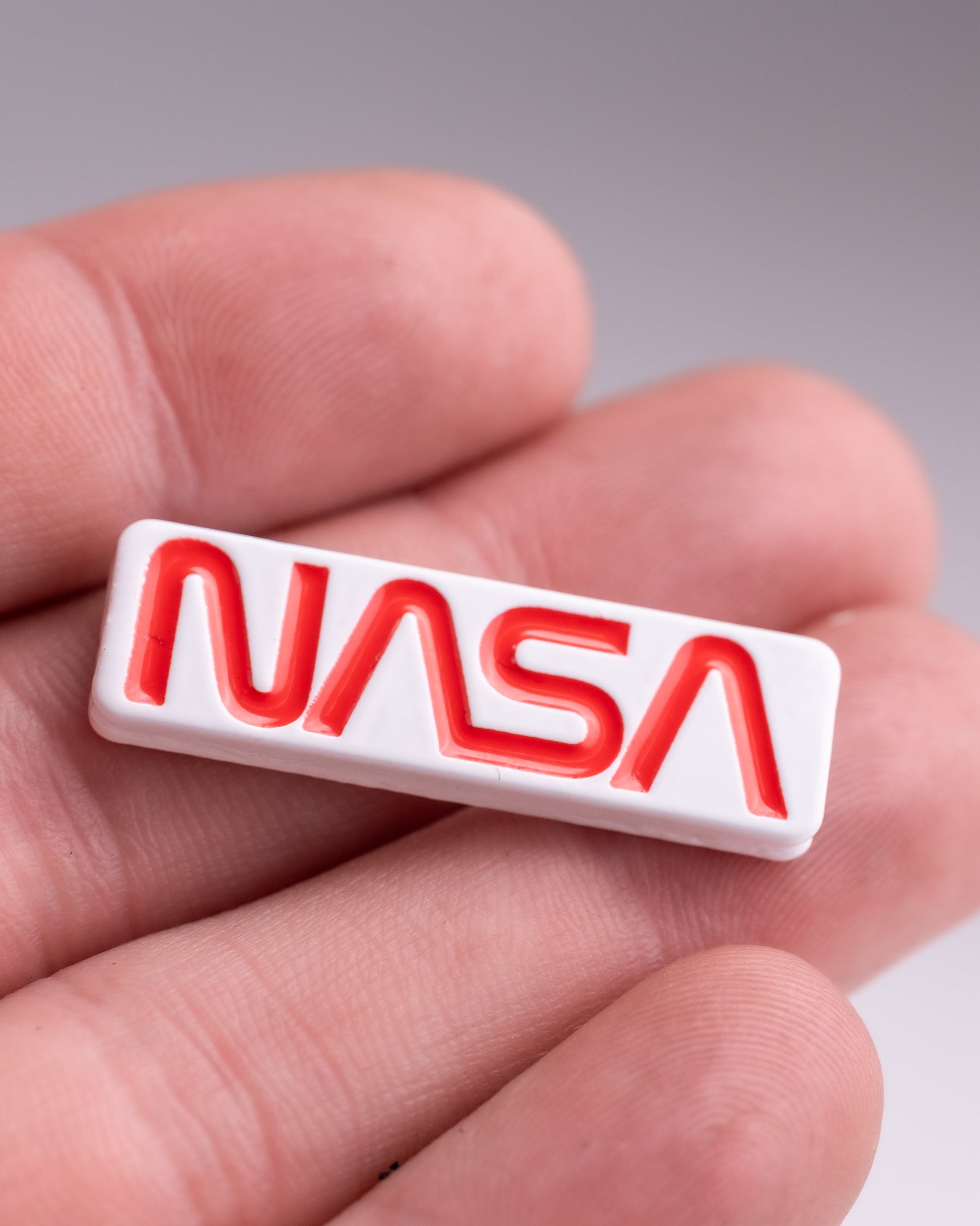 A person holds the NASA "Worm" Ball Marker by Pins and Aces, a small rectangular white marker with the bold red NASA worm logo in enamel. The blurred background emphasizes the marker and the person's fingers.
