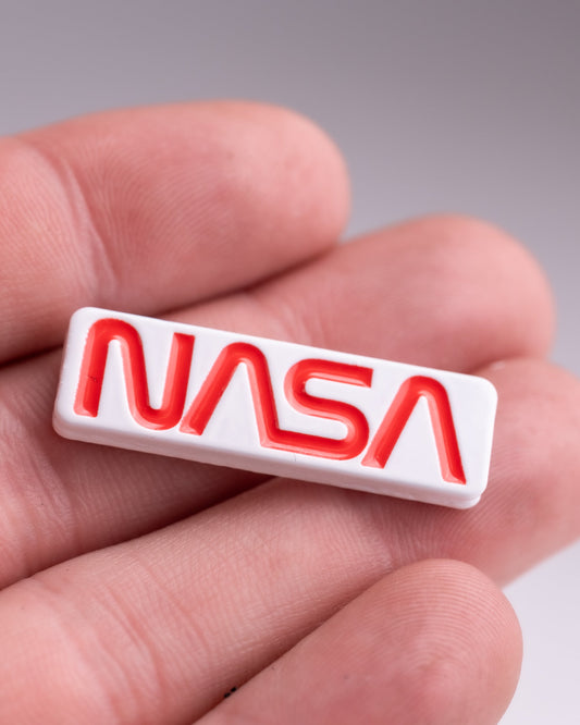 A person holds the NASA "Worm" Ball Marker by Pins and Aces, a small rectangular white marker with the bold red NASA worm logo in enamel. The blurred background emphasizes the marker and the person's fingers.