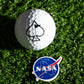 A Pins and Aces NASA "Meatball" Ball Marker with the iconic logo sits on artificial green grass next to a golf ball marked with a black spade symbol.
