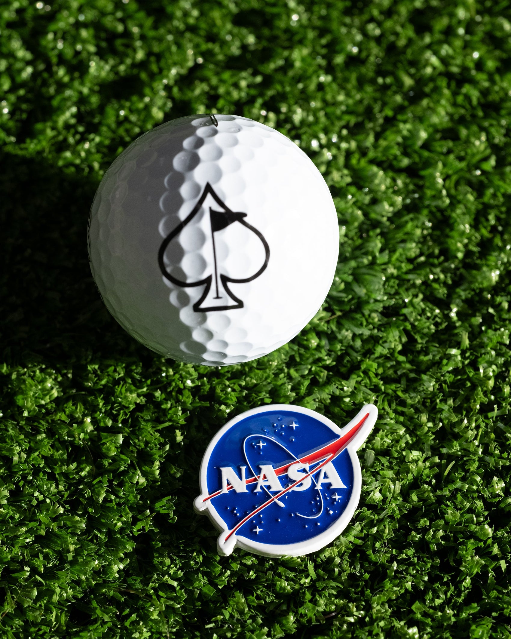 A Pins and Aces NASA "Meatball" Ball Marker with the iconic logo sits on artificial green grass next to a golf ball marked with a black spade symbol.