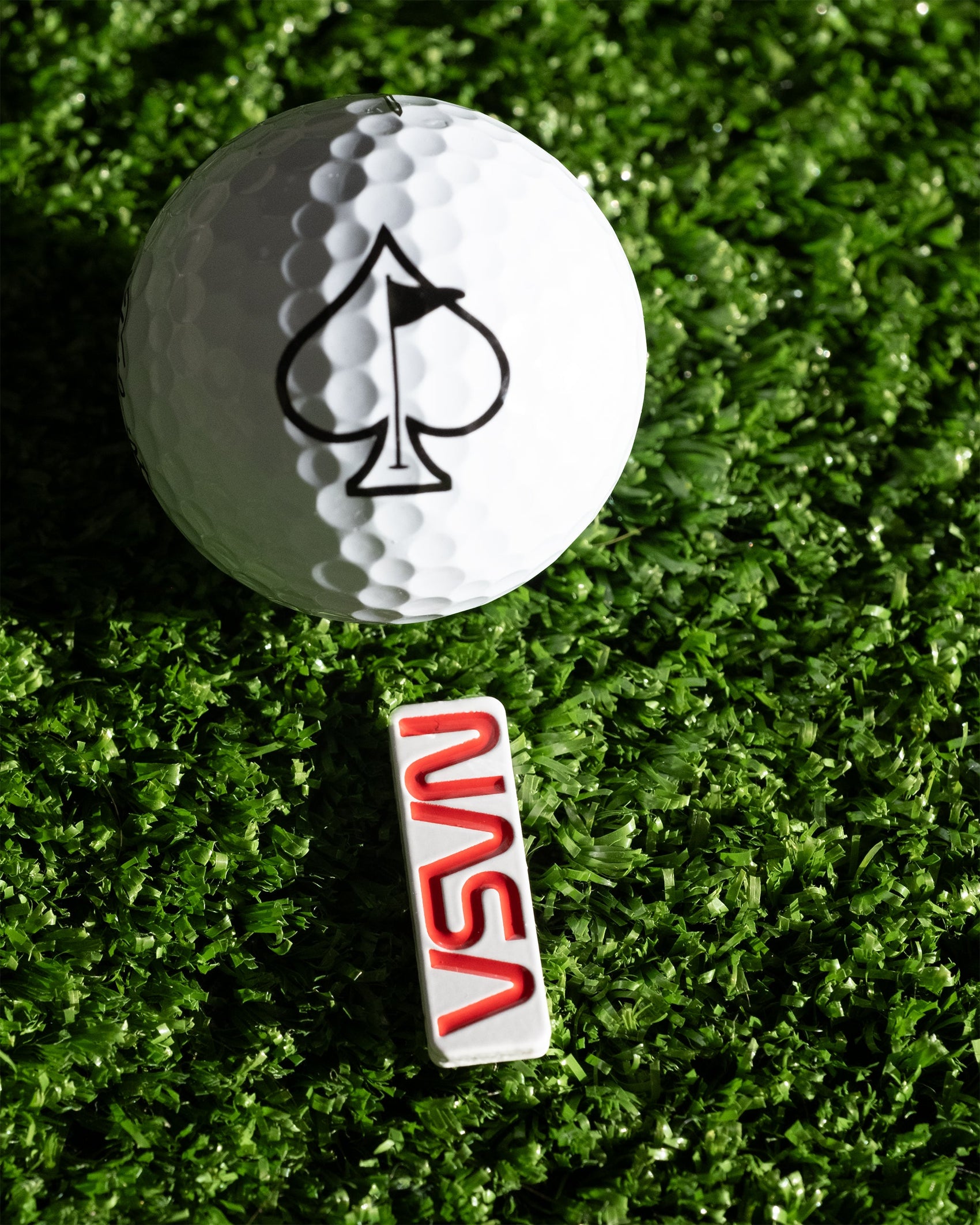 A white golf ball with a black spade sits on green turf above the Pins and Aces NASA "Worm" Ball Marker, styled as an enamel NASA pin.