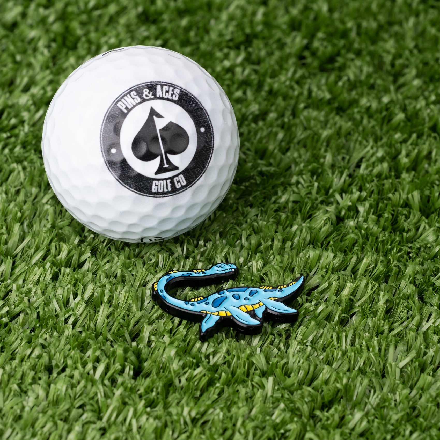 A white golf ball with the Pins and Aces logo and the Nessie Ball Marker, featuring blue enamel, sit on green artificial grass.