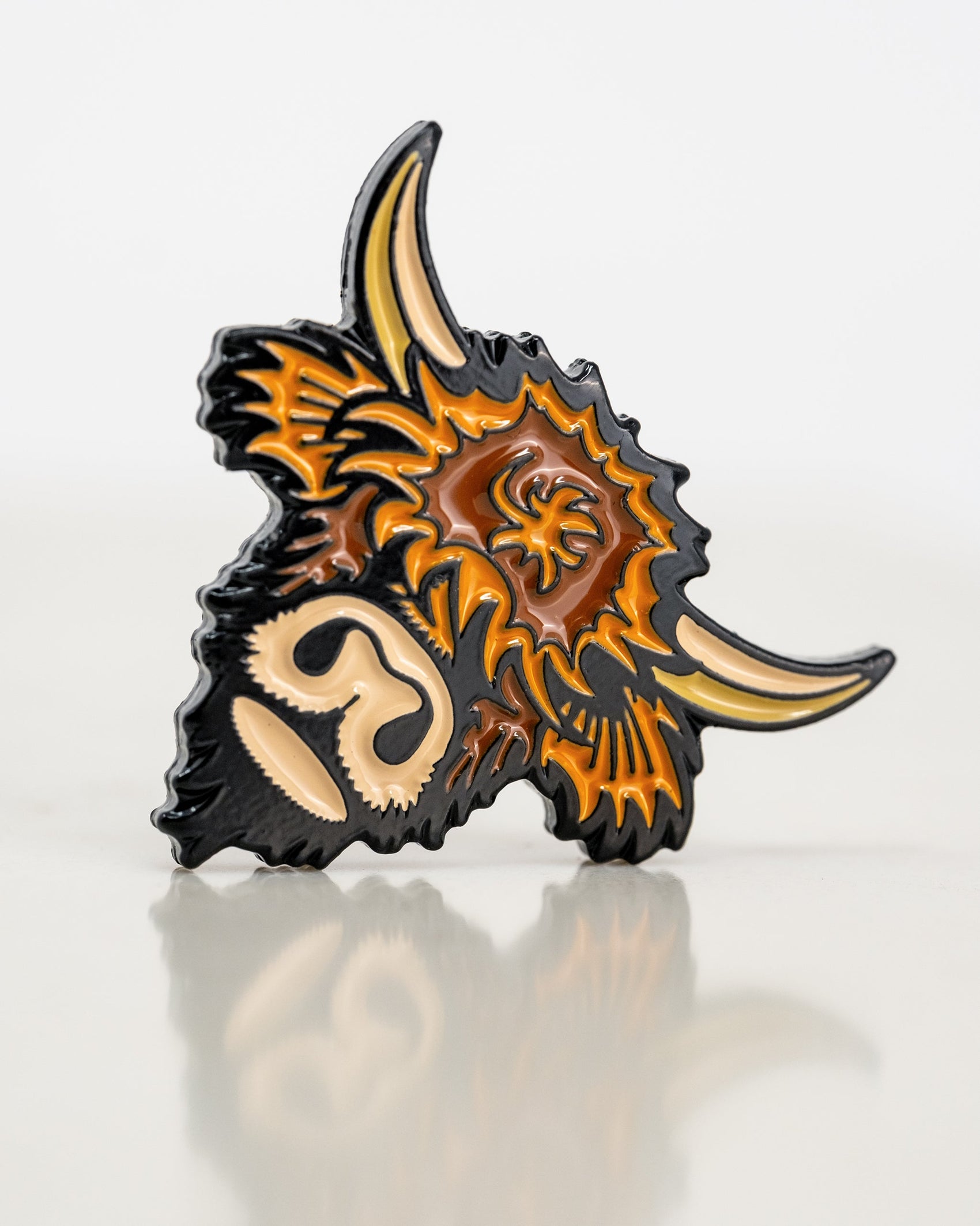 The Highland Cow Ball Marker by Pins and Aces is a cast metal enamel pin shaped like a Highland cow's head, featuring orange, brown, and cream enamel with black outlines, displayed on a reflective white surface.