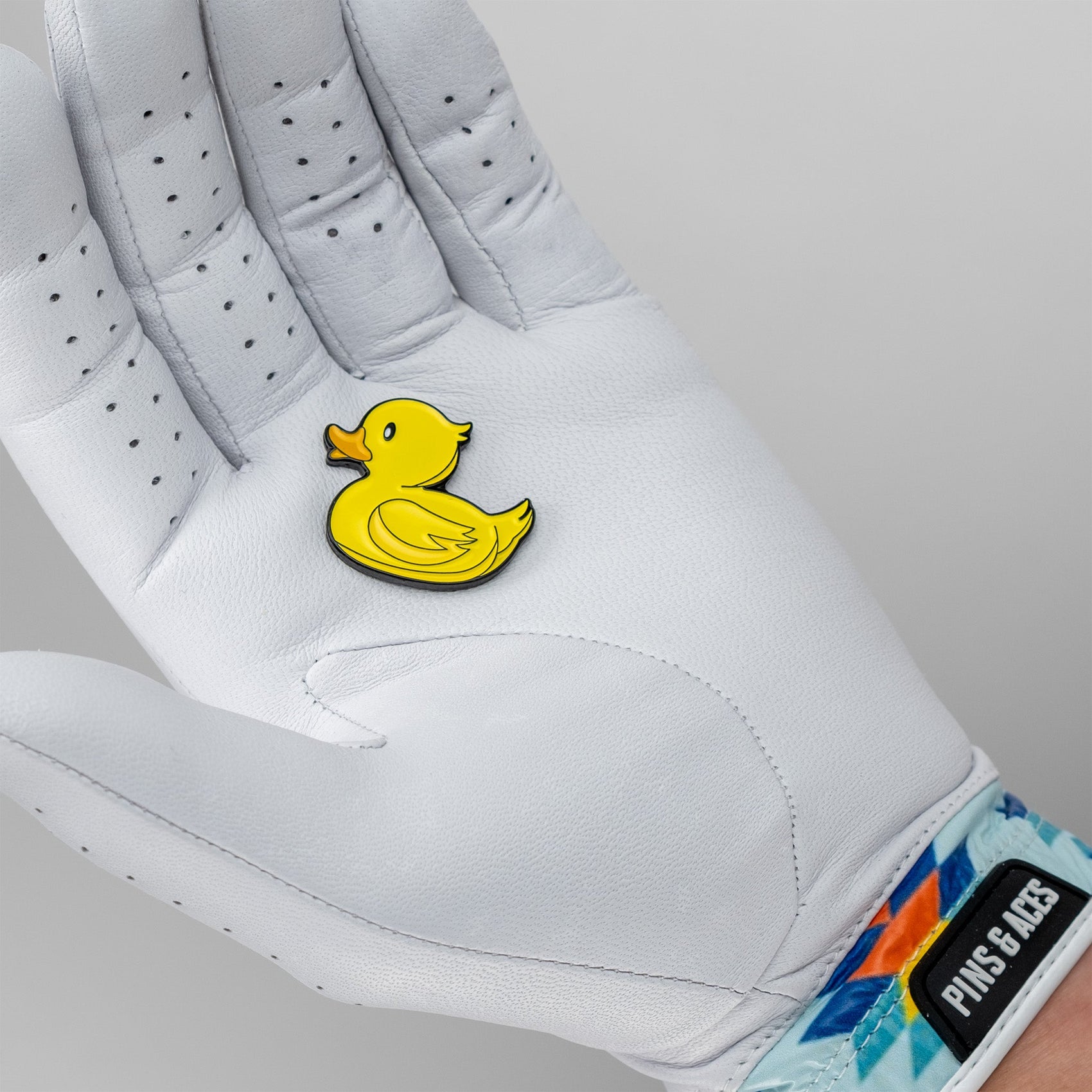 A person wearing a white golf glove with a colorful, geometric wristband holds the Pins and Aces Rubber Ducky Ball Marker in their palm.