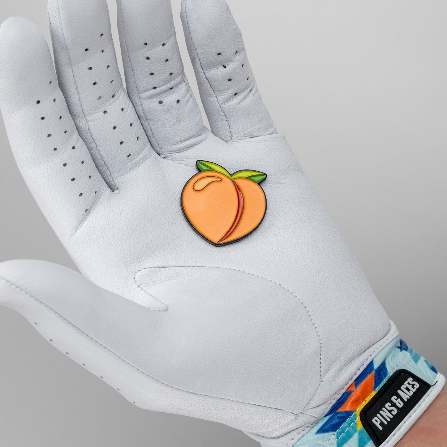 A person wearing a white golf glove with colorful geometric patterns on the wrist holds the Pins and Aces Peach Ball Marker shaped like a peach with green leaves.