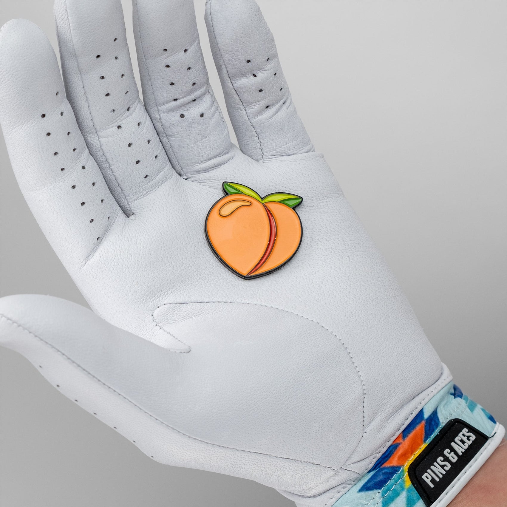 A person wearing a white golf glove with colorful geometric patterns on the wrist holds the Pins and Aces Peach Ball Marker shaped like a peach with green leaves.