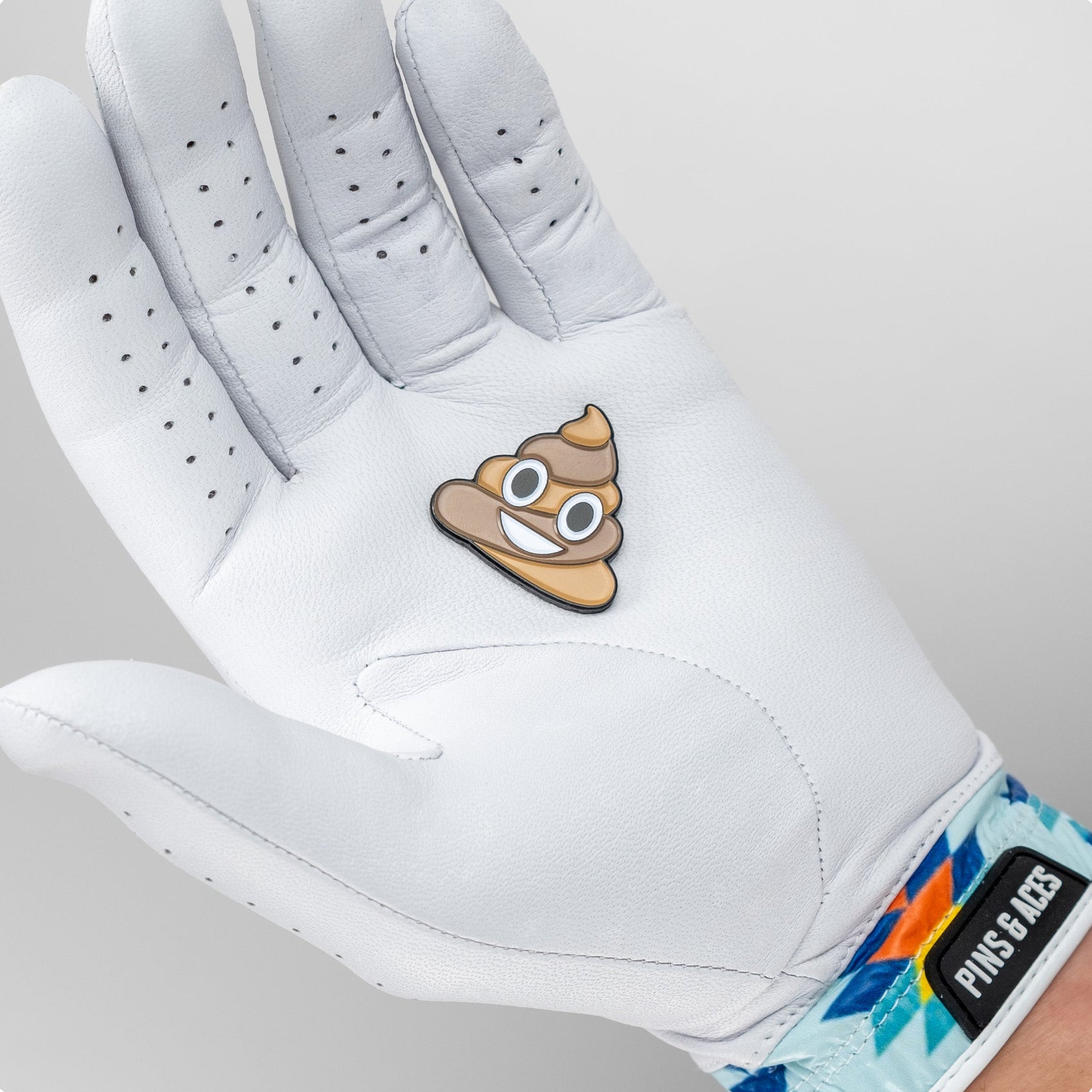 A hand wearing a white golf glove holds the Pins and Aces Poop Ball Marker, an enamel finish ball marker shaped like a smiling poop emoji, with a colorful wristband on the wrist.