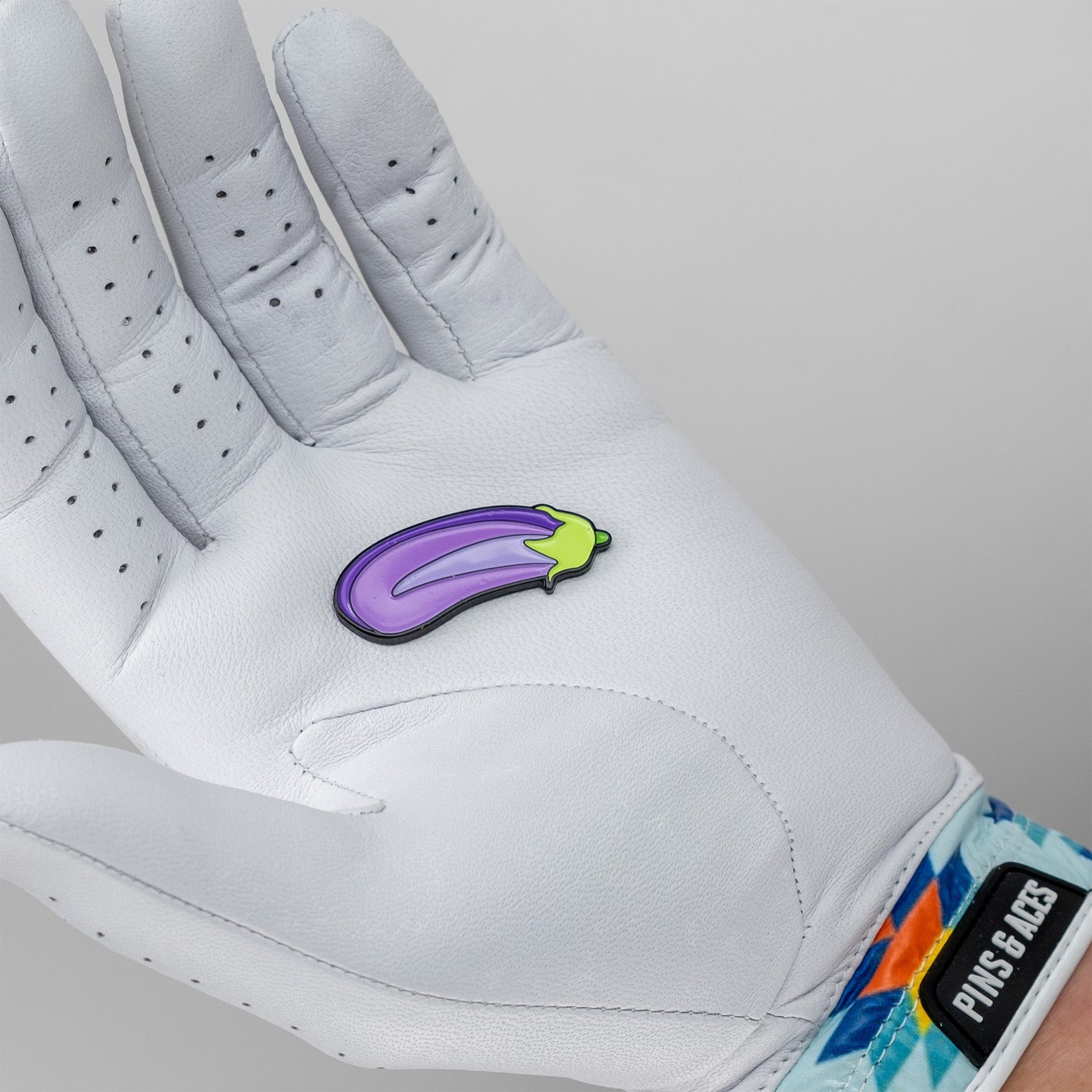 A white-gloved hand holds the Pins and Aces Eggplant Ball Marker, a small purple enamel golf marker shaped like an eggplant. The glove features perforations and a colorful wrist strap with the brand name. Plain gray background.