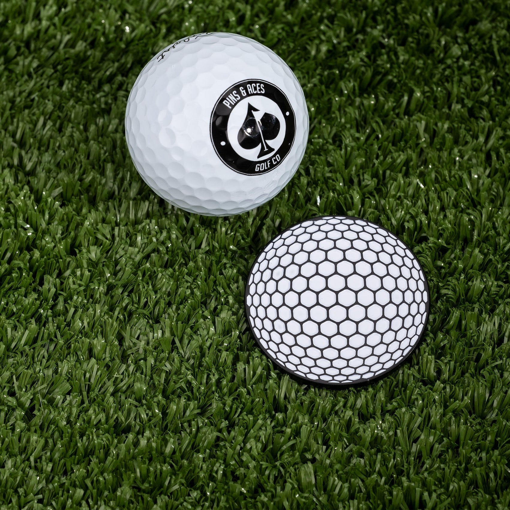 Two golf balls on artificial grass—one with classic dimples and a “Pins and Aces” logo, the other featuring hexagon patterns—make the ideal showcase for stylish Golf Ball Ball Marker accessories by Pins and Aces.