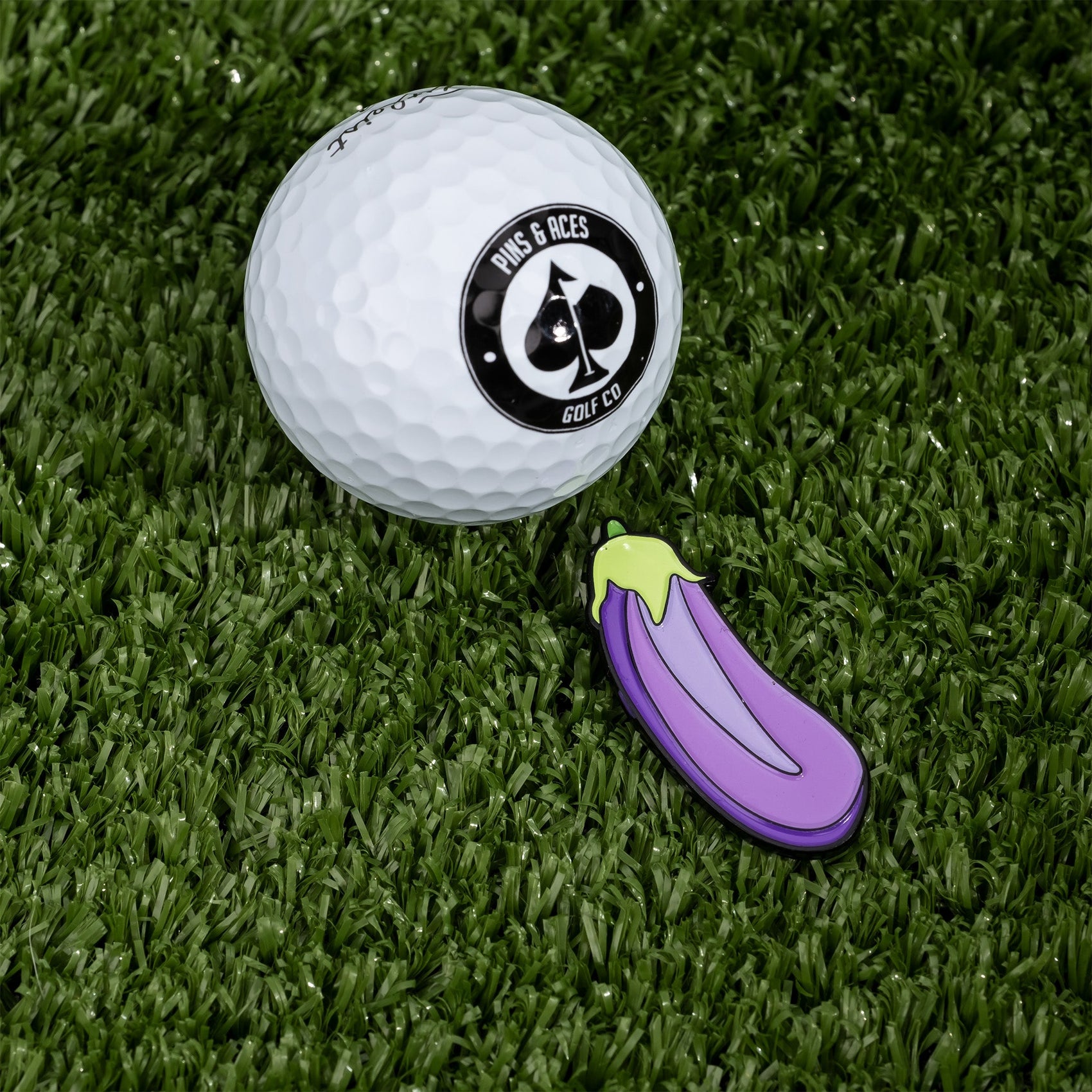 A white golf ball with a Pins and Aces logo rests on grass next to an Eggplant Ball Marker, shaped like a purple eggplant.