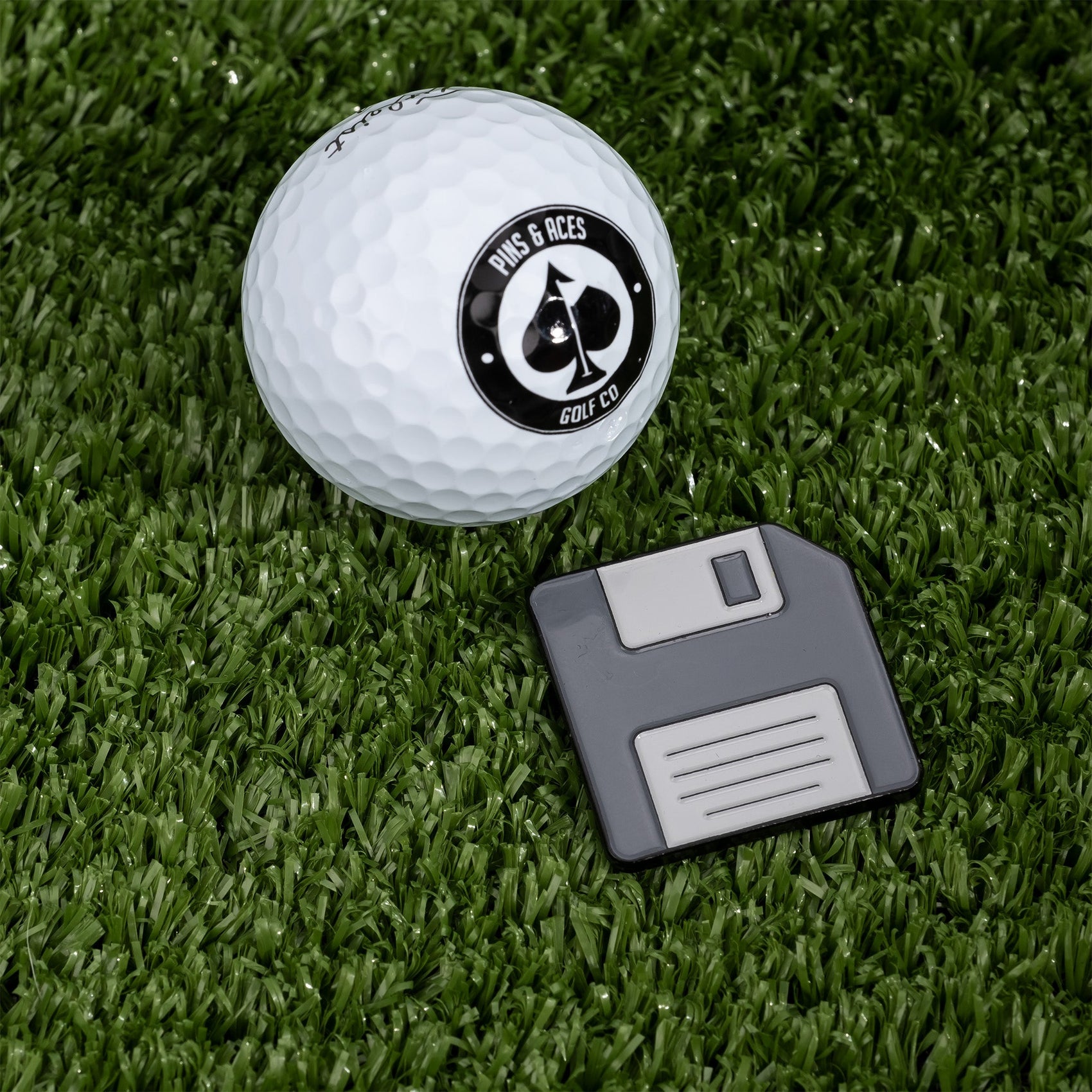 A Pins and Aces golf ball rests on artificial grass beside a gray Floppy Disc Ball Marker, a magnetic marker shaped like a classic floppy disk.